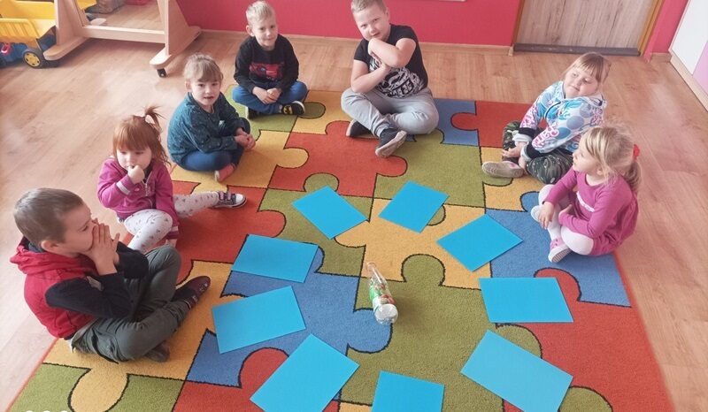 Sala przedszkolna. Dzieci siedzą  w półokręgu na dywanie. Pośrodku dywanu ułożone są w kształcie koła niebieskie kartki . W środku leży plastikowa butelka.