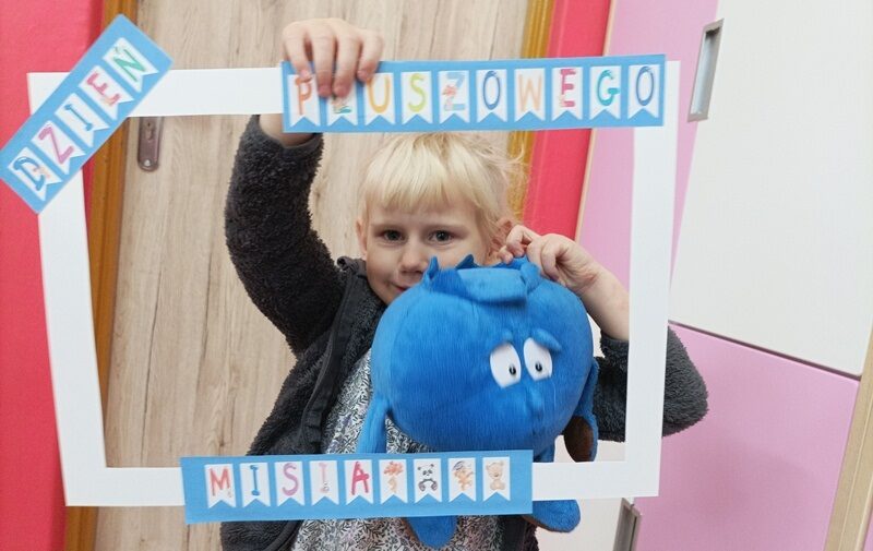 Sala przedszkolna. Dziewczynka trzyma ramkę z kolorowym napisem:  dzień pluszowego misia. W drugiej ręce trzyma niebieską pluszową zabawkę.