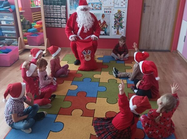 Sala przedszkolna. Na dywanie siedzą dzieci i podnoszą do góry ręce. Przed nimi na krzesełku siedzi Mikołaj.