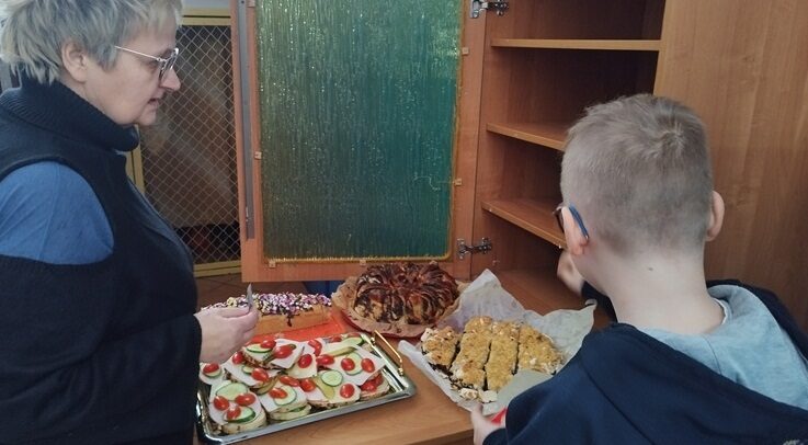 Korytarz szkolny. Nauczycielka i uczeń stoją przy szafie. Na stoliku stoją talerze z ciastem i kanapkami.