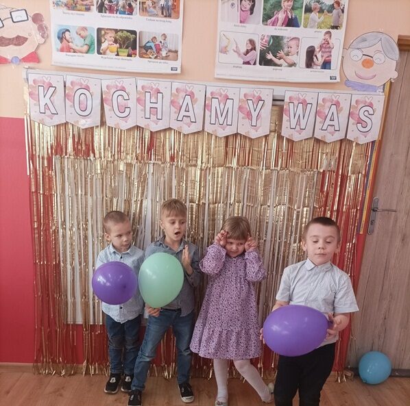 Sala przedszkolna. Czworo dzieci z balonikami stoi przy ścianie pod napisem: Kochamy was.