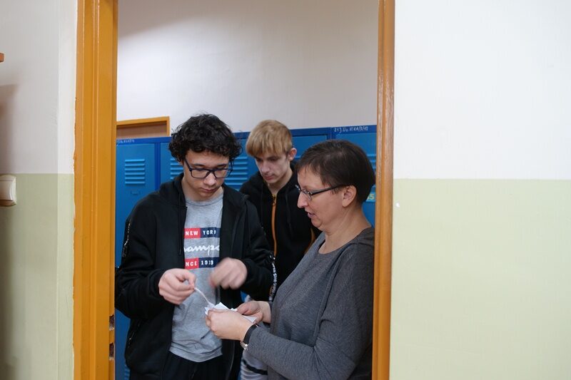 Wejście do sali dydaktycznej. Uczniowie stoją przed wejściem do sali. Jeden z uczniów losuje kartkę.