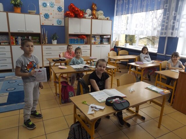 Sala dydaktyczna. Uczniowie siedzą przy stolikach. Jeden uczeń stoi i trzyma plakat.