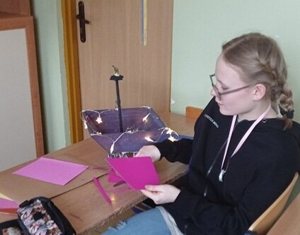 Sala lekcyjna. Uczennica siedzi przy ławce i wycina fioletowe figury z papieru.