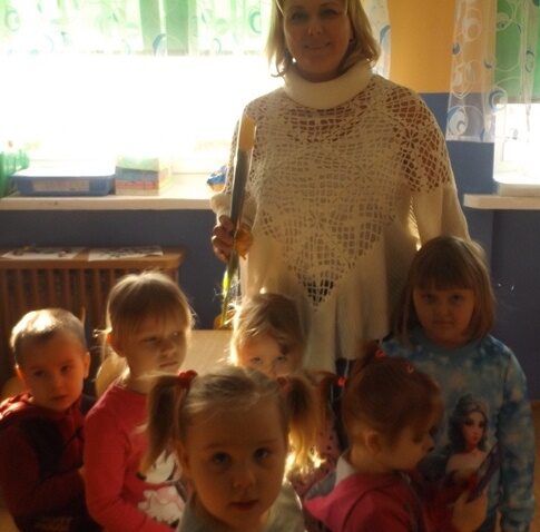 Sala przedszkolna. Nauczycielka z kwiatkiem stoi z grupą dzieci.