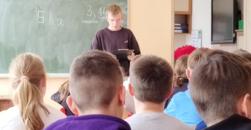 Sala dydaktyczna. Uczeń stoi przy tablicy i czyta tekst. Przed nim na krzesłach siedzą uczniowie.