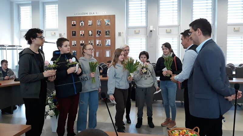 Sala konferencyjna Urzędu Miejskiego w Olecku. Uczniowie z kwiatami i stroikami stoją przed pracownikami urzędu.
