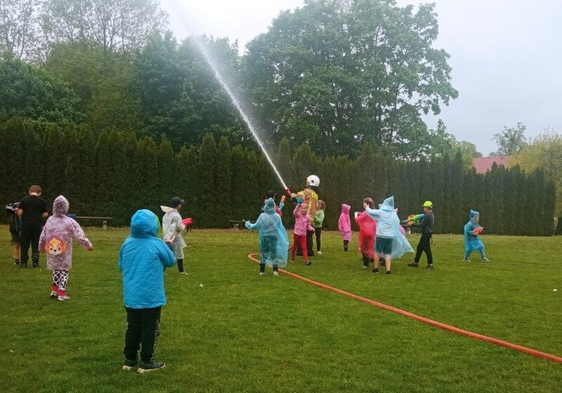 Bosko szkolne. Dzieci stoją na boisku. Strażak prezentuje lanie wody z węża.
