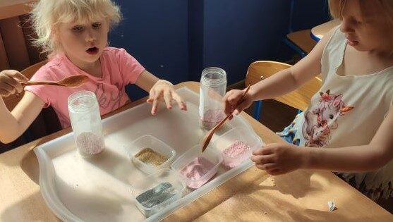 Sala przedszkolna. Dwie dziewczynki siedzą przy stoliku i wsypują kolorowy piasek do słoików.