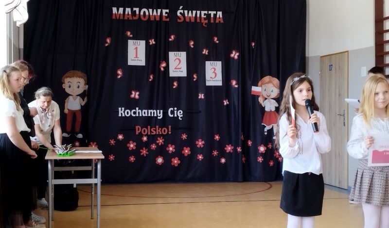 Korytarz szkolny. Napis na kotarze: majowe święta - 1, 2, 3. Kochamy Cię, Polsko! Rysunki dzieci i papierowe biało-czerwone kwiatki. Po lewej stronie na ławce leżą cymbałki. Po prawej stronie stoją dwie dziewczynki i deklamują wiersze.