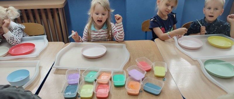 Sala przedszkolna. Dzieci siedzą przy stolikach. Na stolikach materiały do malowania.