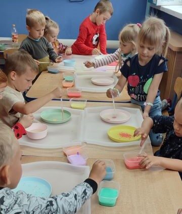 Sala przedszkolna. Dzieci siedzą przy stolikach. Na stolikach materiały do malowania. Dzieci maczają pędzelki w kolorowym mleku.