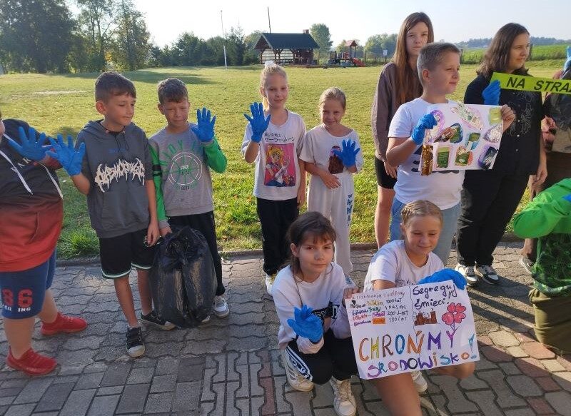 Posesja szkolna. Uczniowie w niebieskich rękawiczkach stoją obok siebie. Dwóch uczniów trzyma plakaty.