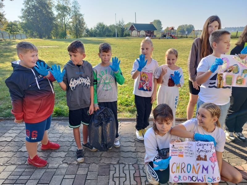 Posesja szkolna. Uczniowie w niebieskich rękawiczkach stoją obok siebie. Dwóch uczniów trzyma plakaty.