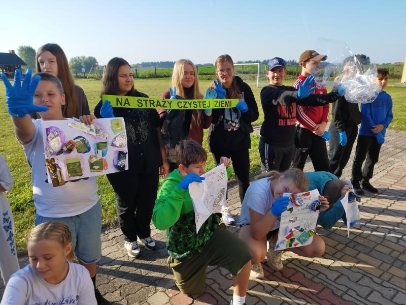 Posesja szkolna. Uczniowie w niebieskich rękawiczkach stoją obok siebie. Uczniowie trzymają plakaty i napis: Na straży czystej Ziemi.