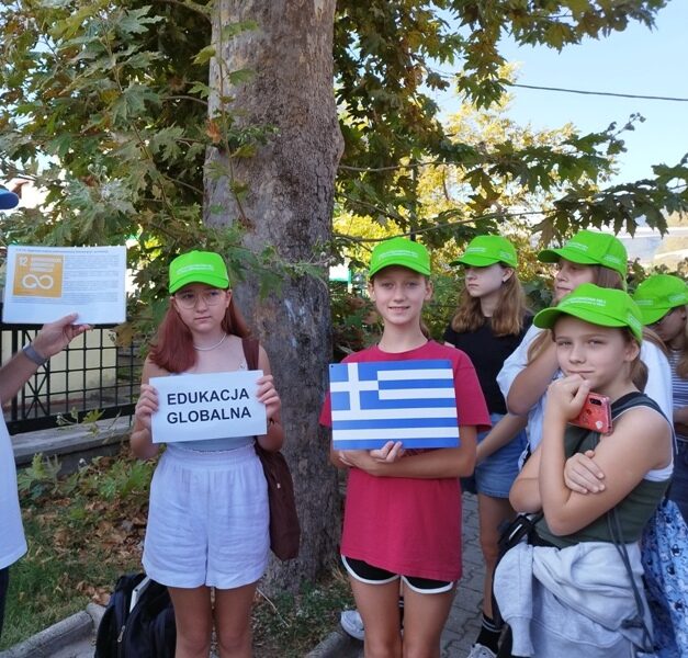 Leptokaria. Nauczyciel stoi przed uczniami. Dwie uczennice trzymają napis Edukacja globalna i małą flagę Grecji.