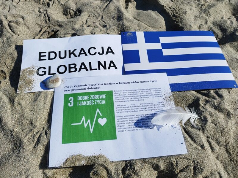 Nei Pori. Na piasku na plaży leżą trzy kartki z napisem Edukacja globalna, cel 3, flaga Grecji.