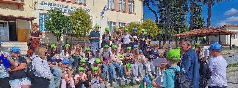 Uczniowie siedzą przed szkołą. Przed nimi stoi nauczyciel.