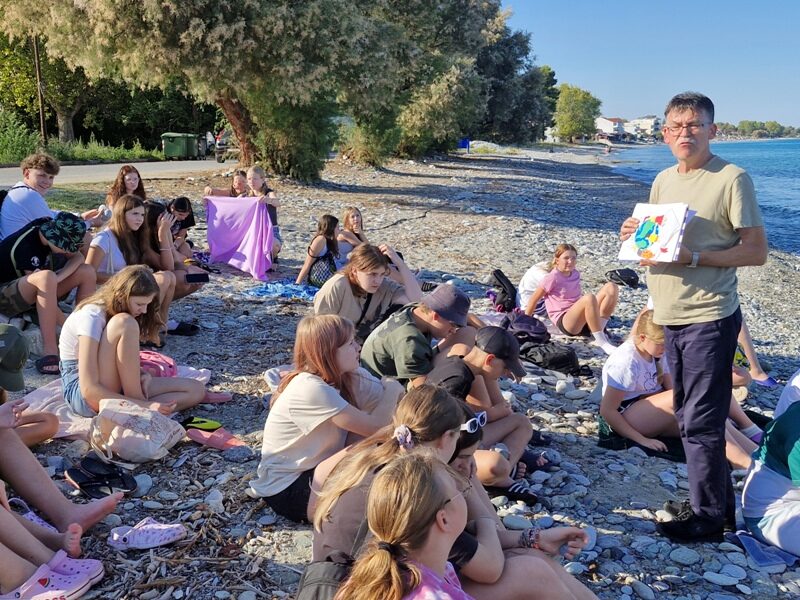 Nad Morzem Egejskim. Uczniowie siedzą na plaży. Przed nimi stoi nauczyciel.