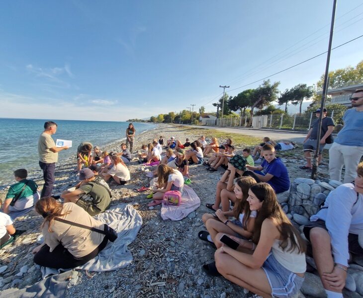 Nad Morzem Egejskim. Uczniowie siedzą na plaży. Przed nimi stoi nauczyciel.