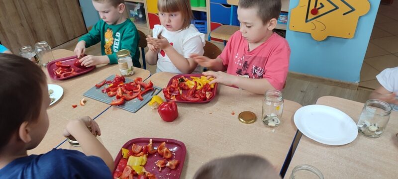 Sala przedszkolna. Dzieci siedzą przy stolikach. Na stolikach znajdują się papryki, talerzyki i słoiki.