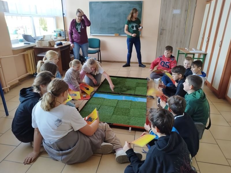 Sala dydaktyczna. Uczniowie siedzą na podłodze przy planszy.