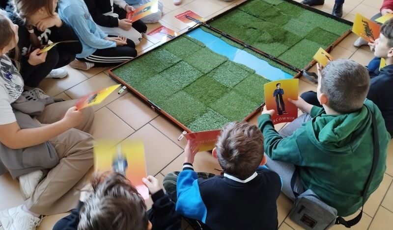 Sala dydaktyczna. Uczniowie siedzą na podłodze przy planszy.